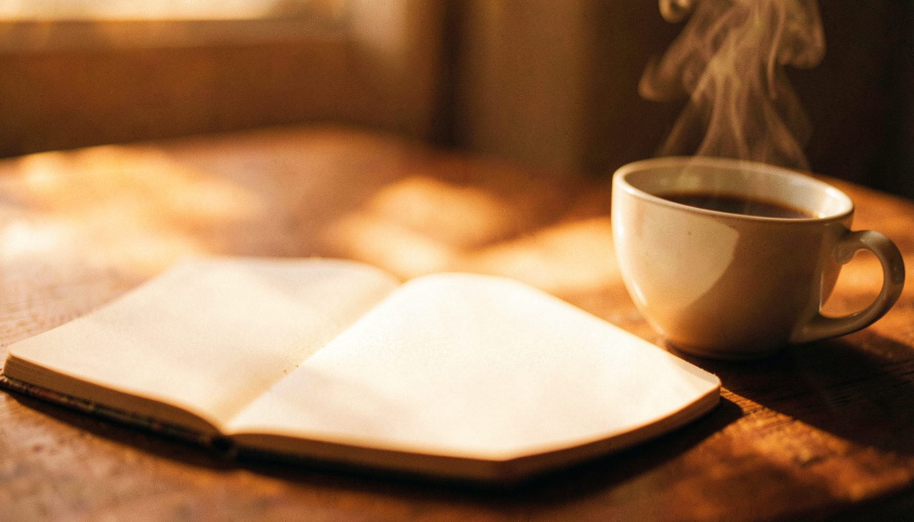 Soft-focus close-up of a blank open notebook on a wooden desk beside a steaming cup of coffee, warm morning light spilling across the scene, cream and brown tones with a calm, reflective mood