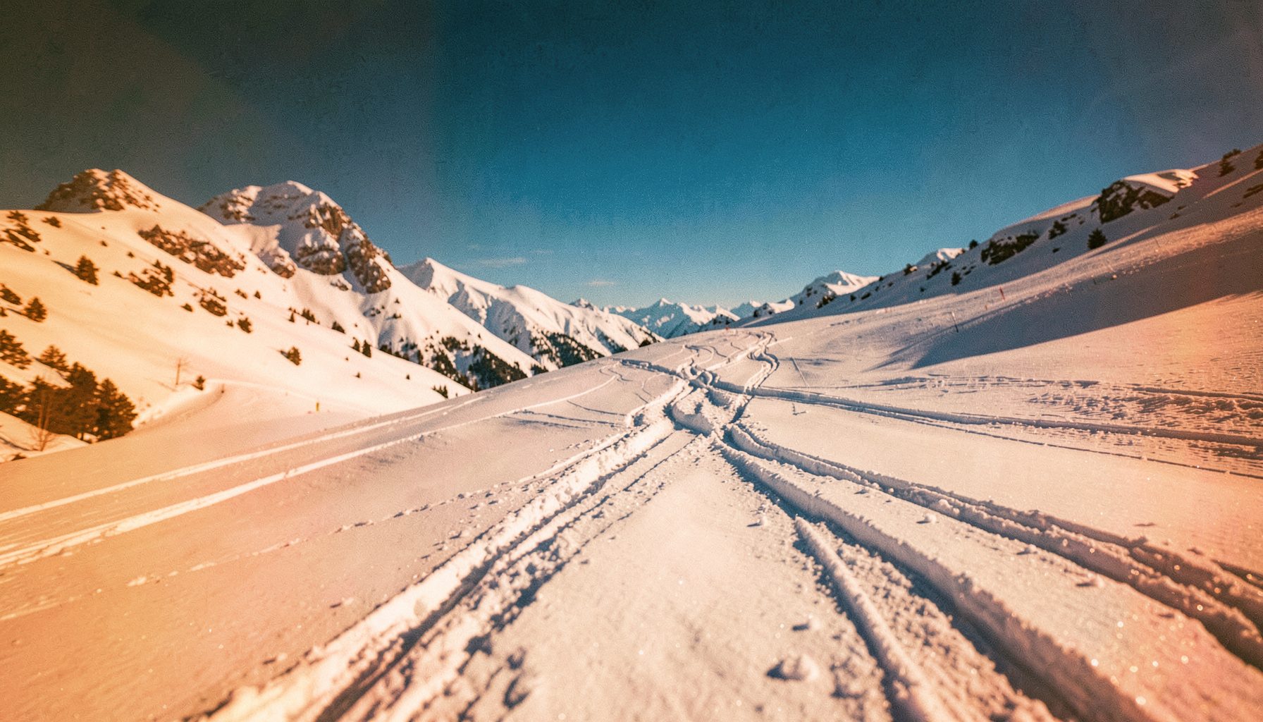 Wide cinematic frame of a snowy mountain slope under bright sunlight with ski tracks cutting through fresh powder, crisp blue sky above