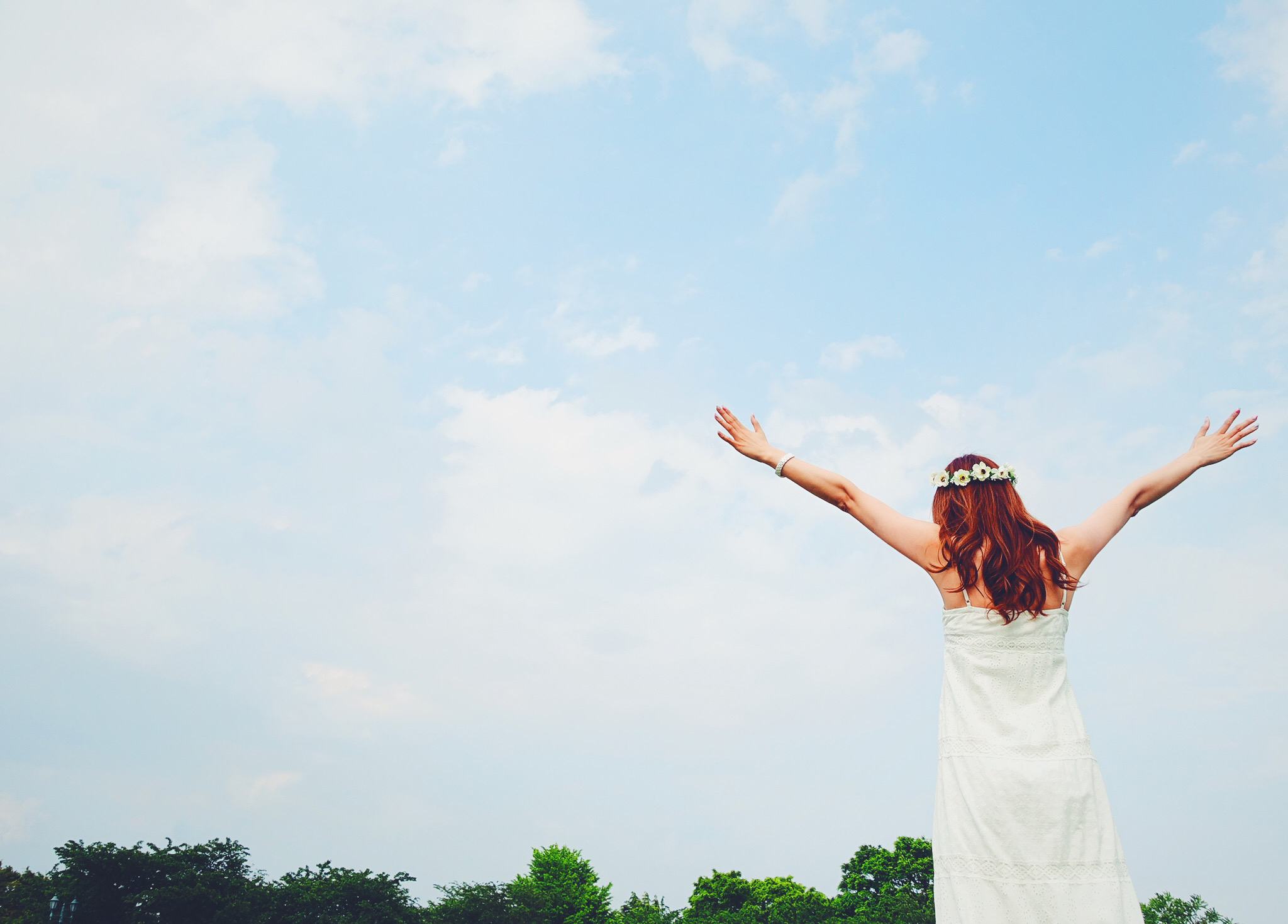 Woman in white dress with flower crown seen from behind, arms raised toward a blue sky with clouds and treeline