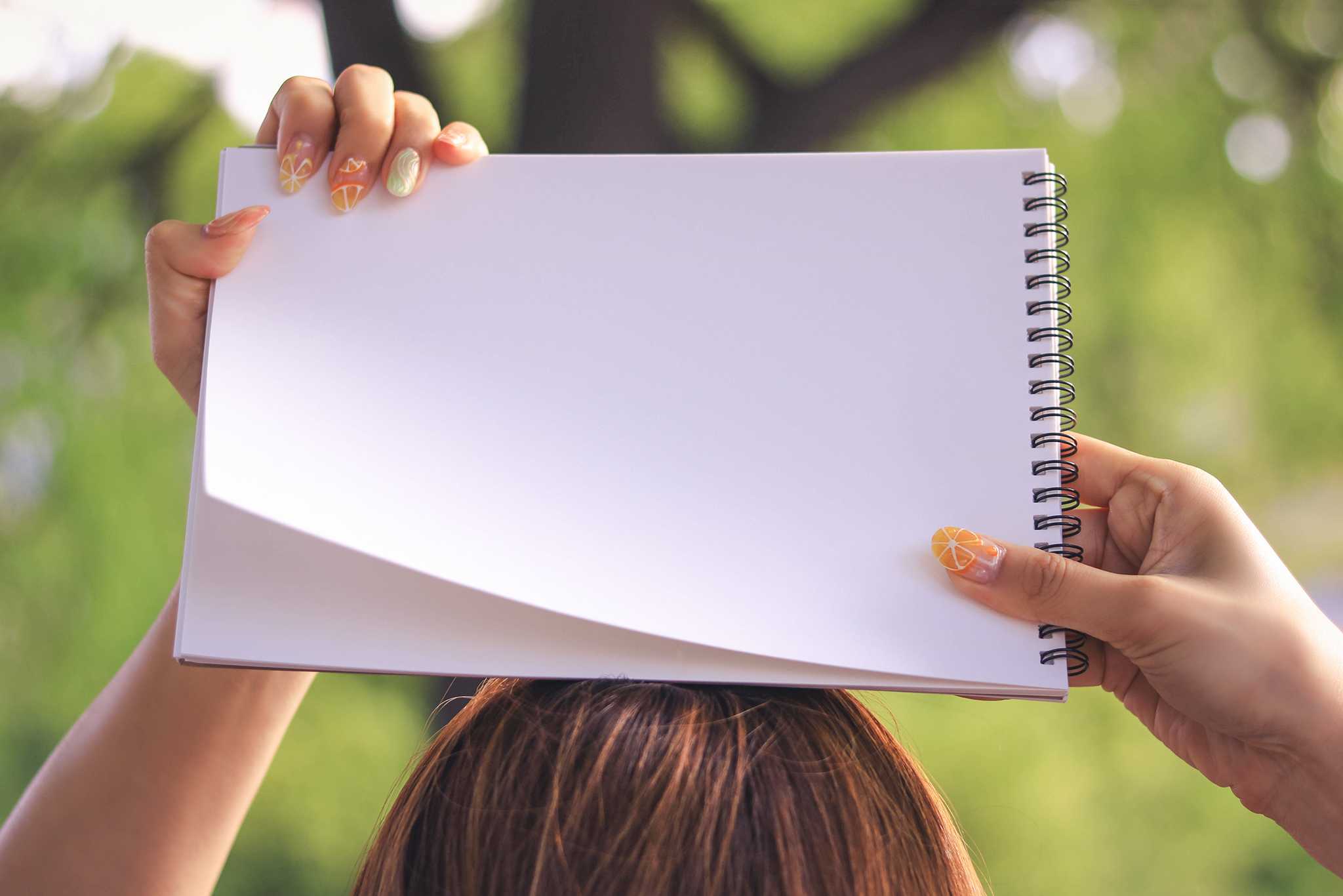 Close-up of hands with orange-patterned nail art holding a blank spiral notebook above a person's head, green foliage background